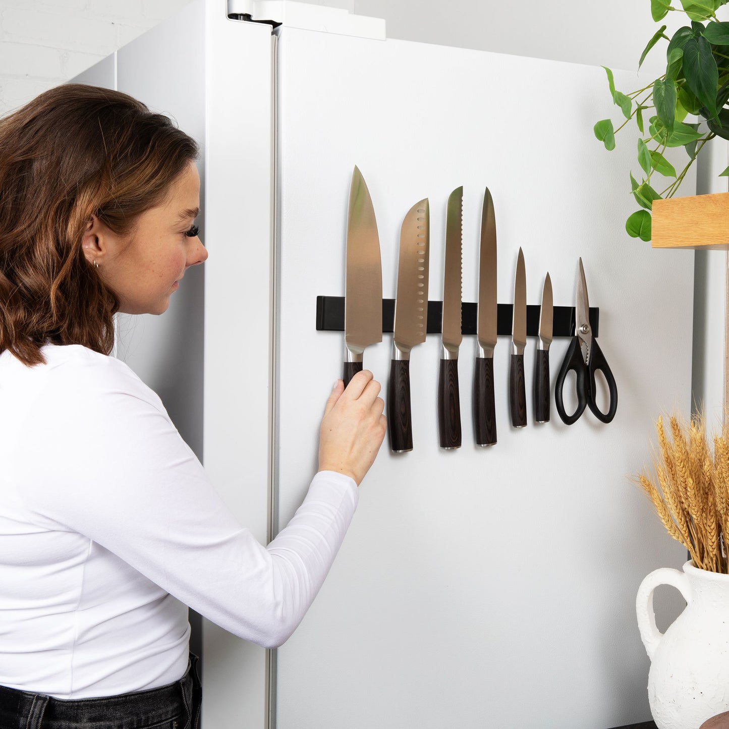 Woman using a magnetic knife strip on a refrigerator with various knives and scissors.
