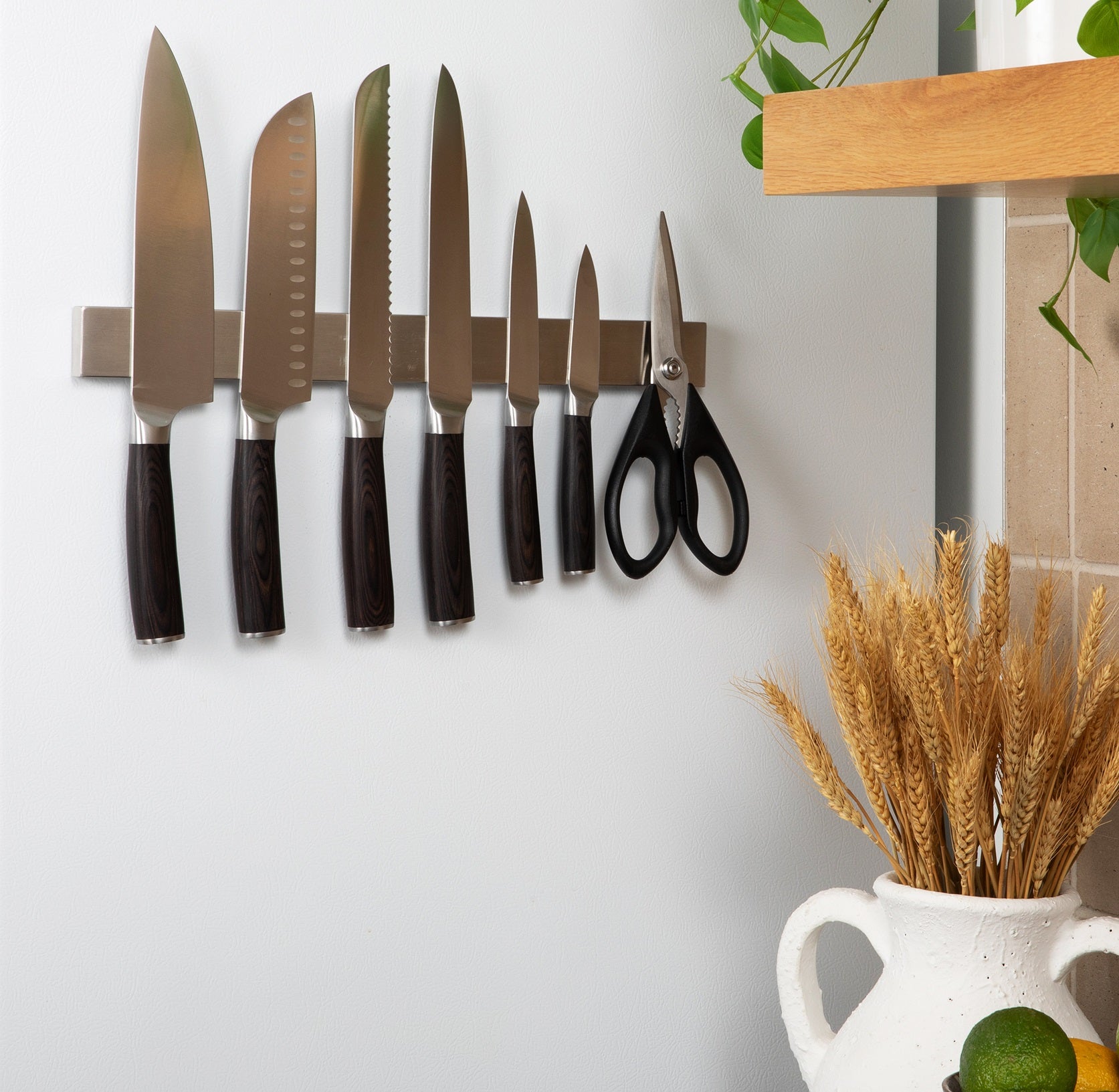 Set of knives and scissors on a a  pampvanna's magnetic strip against a white wall with a vase of wheat in the foreground.